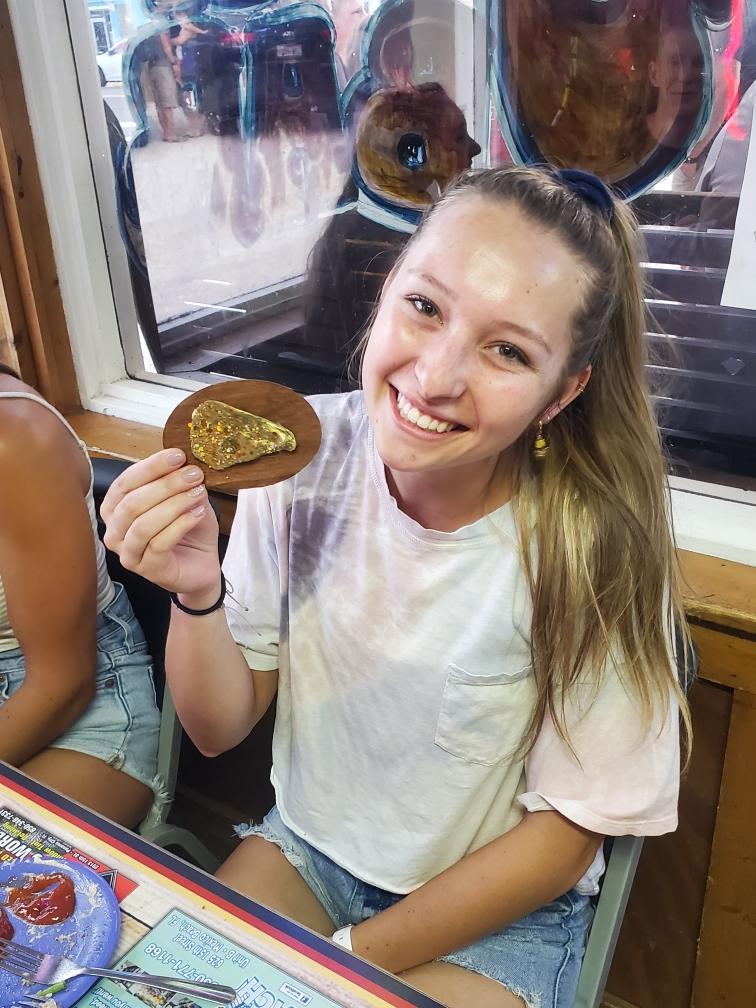 National Oyster Day Shines Bright with the Golden Oyster at Hunt’s in Panama City: A Truly Unforgettable Celebration 7 A young woman smiles while holding a golden oyster at Hunt's Oyster Bar in Panama City, Florida during a National Oyster Day celebration. A festive atmosphere surrounds her with friends and painted windows.