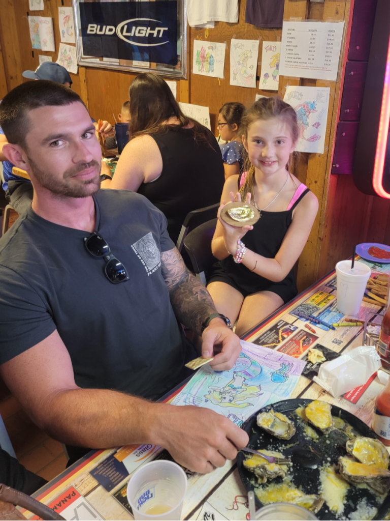 National Oyster Day Shines Bright with the Golden Oyster at Hunt’s in Panama City: A Truly Unforgettable Celebration 5 A family celebrates National Oyster Day at Hunt's Oyster Bar in Panama City, Florida. A man and young girl sit at a table with cheesy oysters, coloring pages, and a golden oyster prize.
