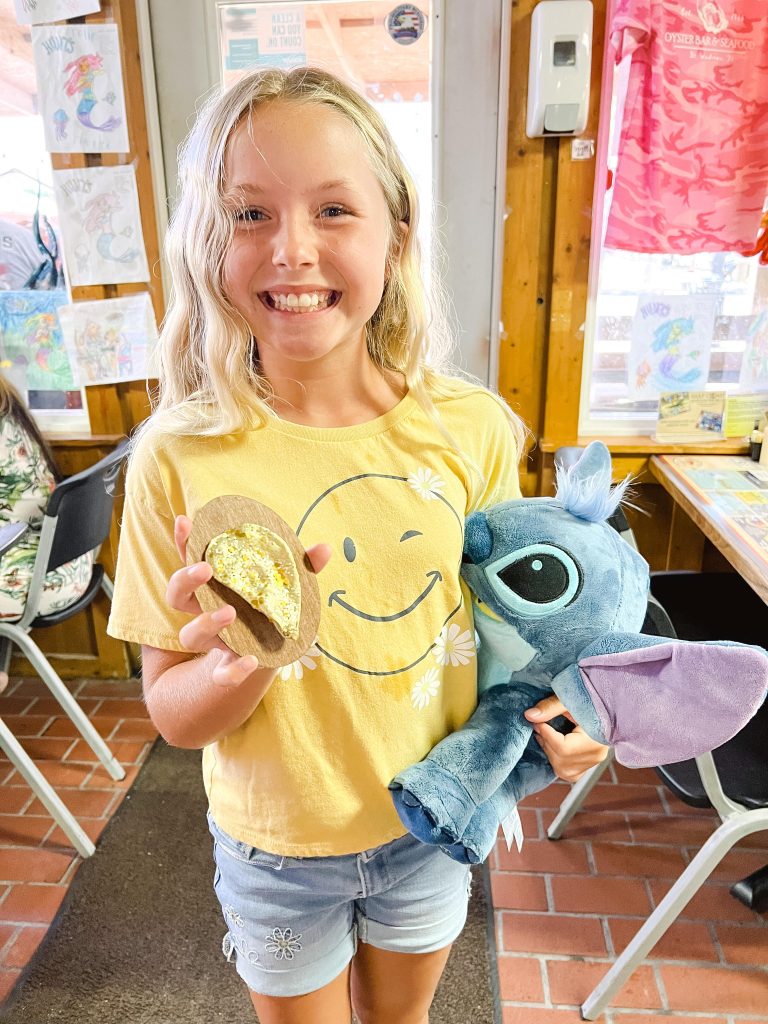 National Oyster Day Shines Bright with the Golden Oyster at Hunt’s in Panama City: A Truly Unforgettable Celebration 6 A smiling girl holding a plush toy and a golden oyster celebrates National Oyster Day at Hunt's Oyster Bar in Panama City, Florida. Behind her, colorful mermaid drawings line the wall.