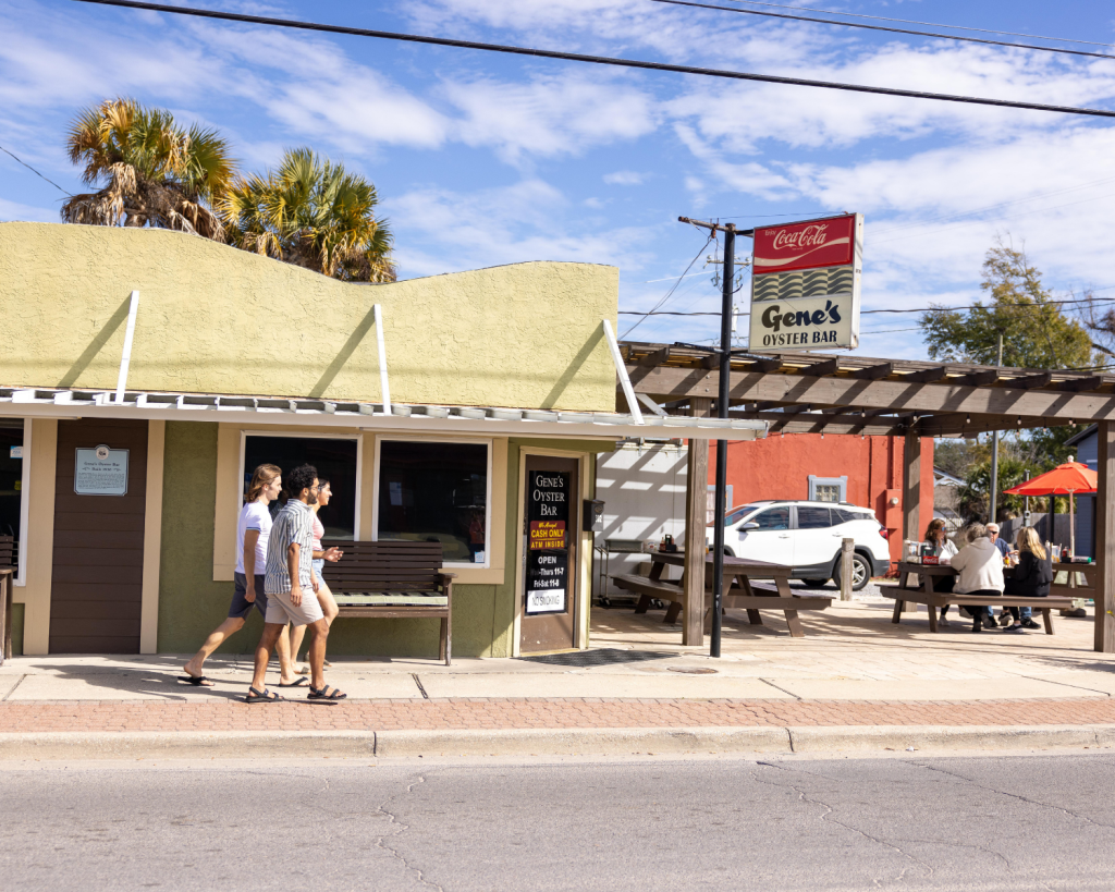 The historic exterior of Gene's Oyster Bar in Panama City, Florida, with visitors strolling by and patrons dining at picnic tables.