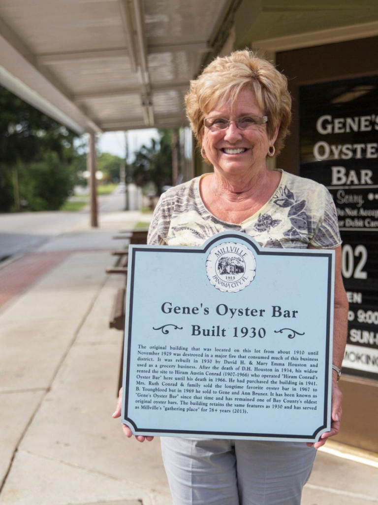 A woman proudly holds the historical marker detailing the legacy of Gene's Oyster Bar in Panama City, Florida, established in 1930.