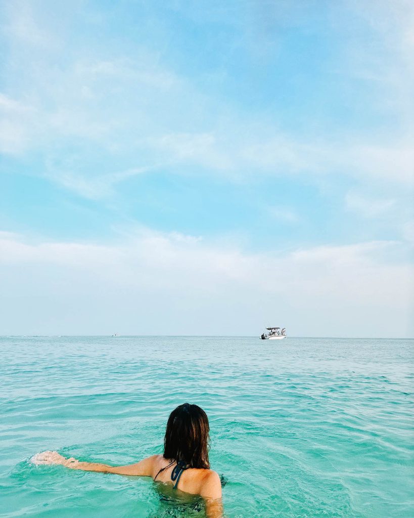 A woman enjoying spring break in Panama City, Florida, floating in the clear turquoise water while gazing at a boat in the distance. The sky is bright and blue, creating a perfect scene for a relaxing and adventurous getaway.