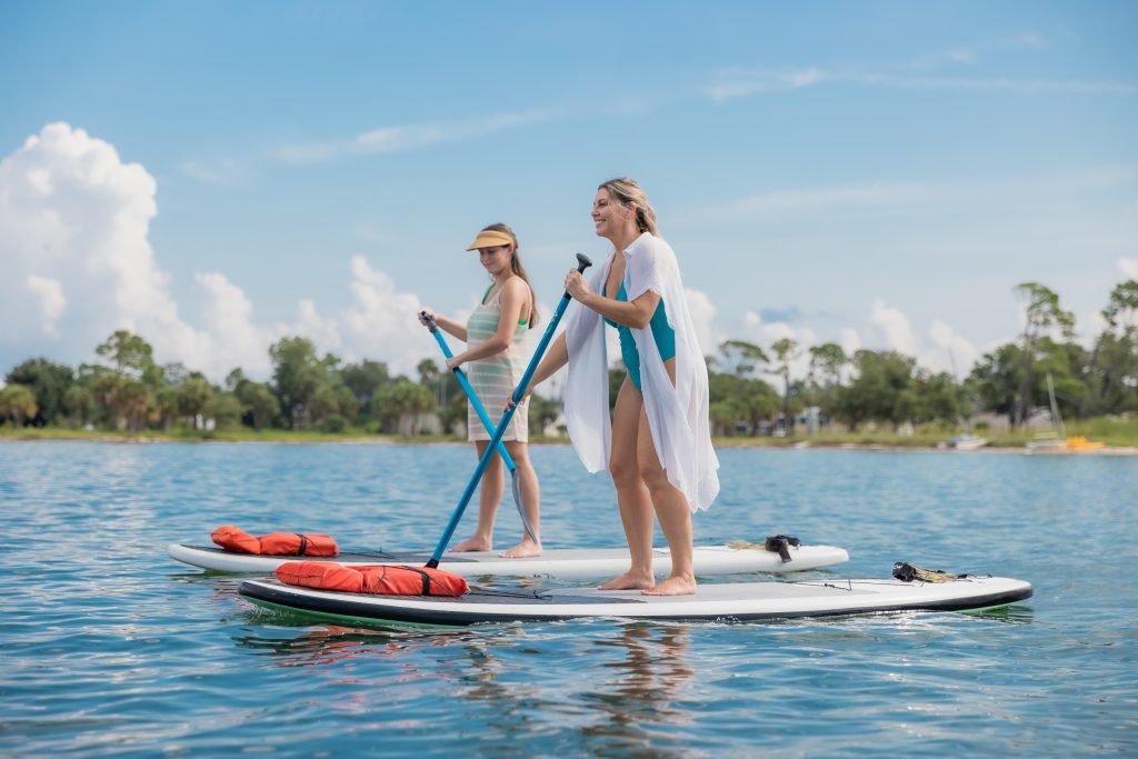 Two women on paddleboards in St. Andrews Bay, surrounded by blue waters, a bright blue sky, and fluffy white clouds—an ideal scene for Florida eco-adventures and sustainable travel in Panama City.