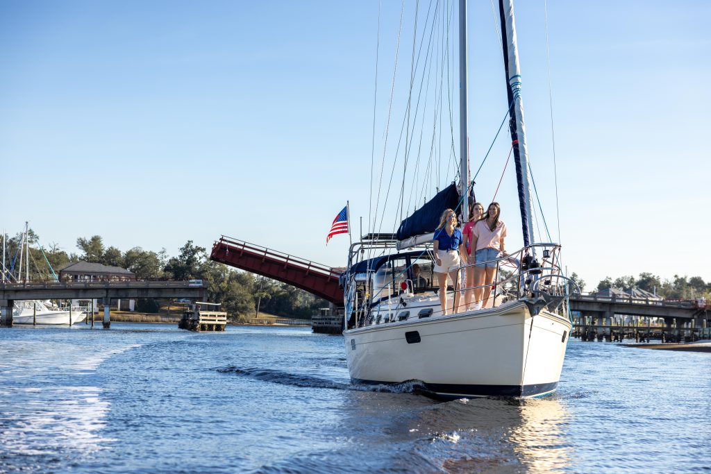 Panama City - Home to World Champions, Historic Marvels, and Rare Natural Wonders in Florida 5 Three girls sailing on St. Andrews Bay with the Tarpon Dock Bridge rising in the background, capturing a scenic moment near one of the historic natural wonders in Florida.