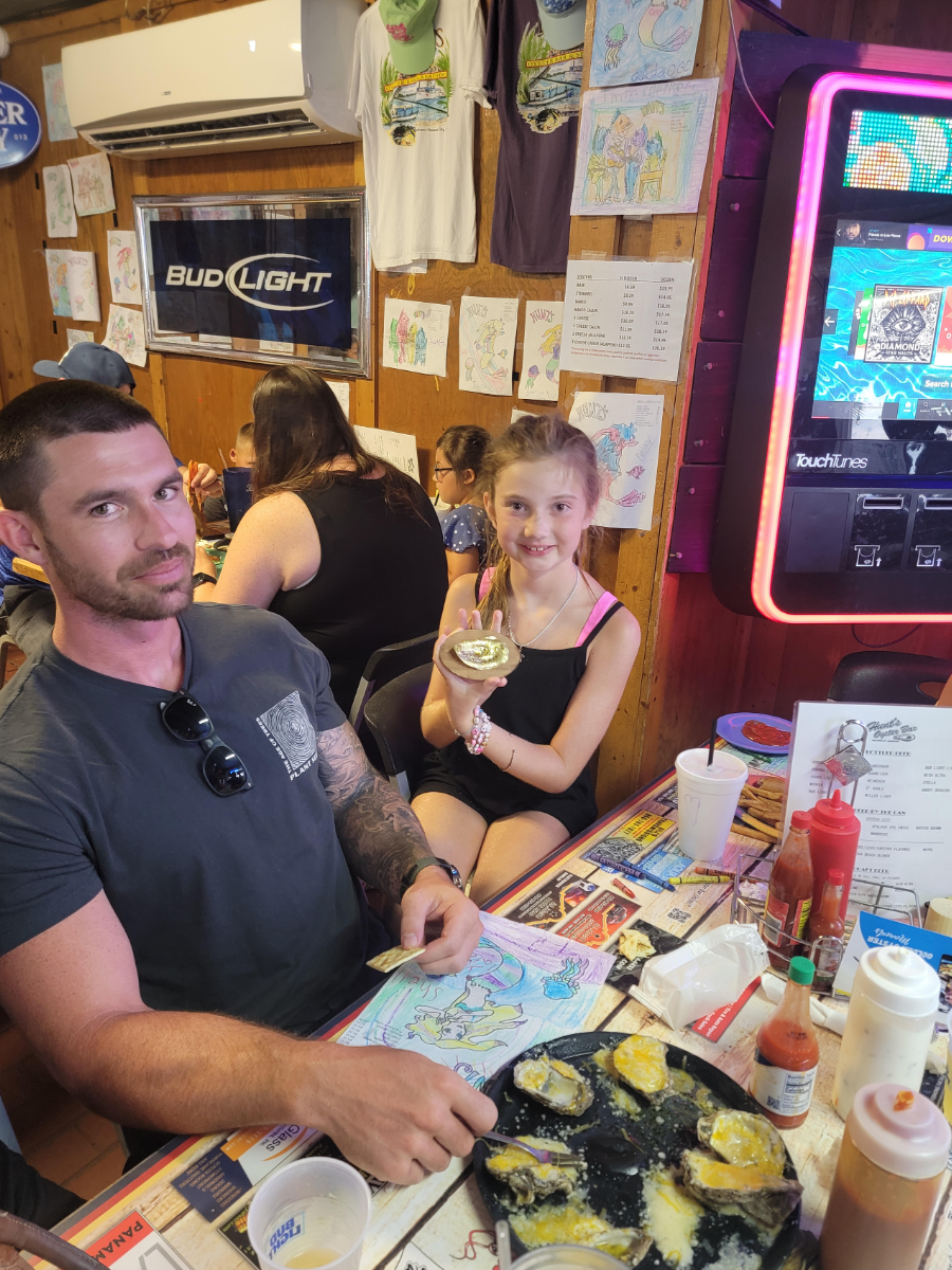 National Oyster Day Shines Bright with the Golden Oyster at Hunt’s Oyster Bar in Panama City, Florida 4 A family celebrates National Oyster Day at Hunt's Oyster Bar in Panama City, Florida. A man and young girl sit at a table with cheesy oysters, coloring pages, and a golden oyster prize.
