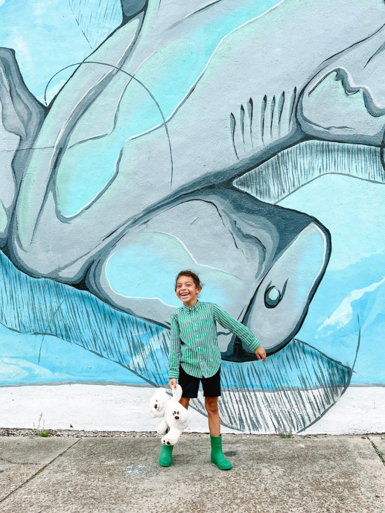 Top 10 Activities for a Family-Friendly Spring Break in Panama City, Florida 7 Child smiling in front of a colorful fish mural on the Panama City Mural Trail during a family-friendly spring break in Panama City.