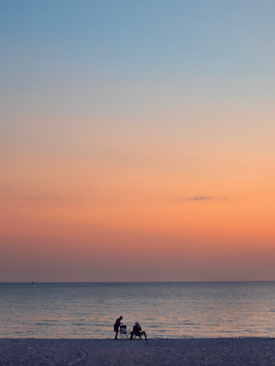 Where to Watch the Best Sunset in Panama City, Florida 3 Visitors relaxing on the beach at St. Andrews State Park beneath a soft orange sunset in Panama City Florida.